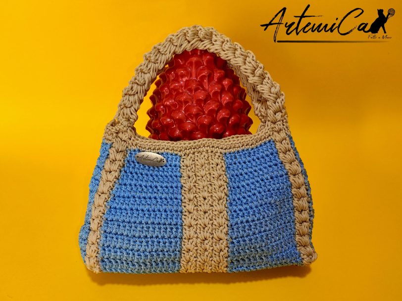 L'immagine mostra una borsa azzurra all'uncinetto con strisce verticali gialle, dotata di un manico coordinato e una chiusura magnetica. Sogno Mediterraneo: il fascino del mare nella tua mano.