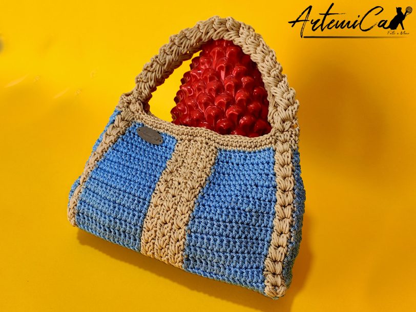 L'immagine mostra una borsa azzurra all'uncinetto con strisce verticali gialle, dotata di un manico coordinato e una chiusura magnetica. Sogno Mediterraneo: il fascino del mare nella tua mano.