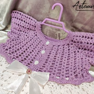 Il Vestitino handmade per bambina su artemicat.live è ritratto in tutta la sua bellezza. La giacchetta a mezza manica in lilla chiaro si abbina splendidamente alla gonnellina bianca, mentre il fiocco decorativo al centro aggiunge un tocco di eleganza. I dettagli artigianali, come i bottoni dorati e il lavoro all'uncinetto, sono chiaramente visibili, evidenziando la qualità e l'amore dedicato alla creazione di questo incantevole capo per bambini.