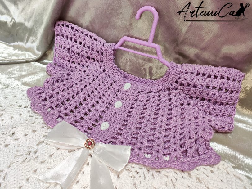 Il Vestitino handmade per bambina su artemicat.live è ritratto in tutta la sua bellezza. La giacchetta a mezza manica in lilla chiaro si abbina splendidamente alla gonnellina bianca, mentre il fiocco decorativo al centro aggiunge un tocco di eleganza. I dettagli artigianali, come i bottoni dorati e il lavoro all'uncinetto, sono chiaramente visibili, evidenziando la qualità e l'amore dedicato alla creazione di questo incantevole capo per bambini.