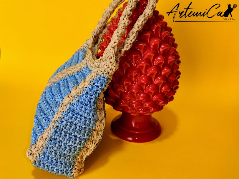 L'immagine mostra una borsa azzurra all'uncinetto con strisce verticali gialle, dotata di un manico coordinato e una chiusura magnetica. Sogno Mediterraneo: il fascino del mare nella tua mano.