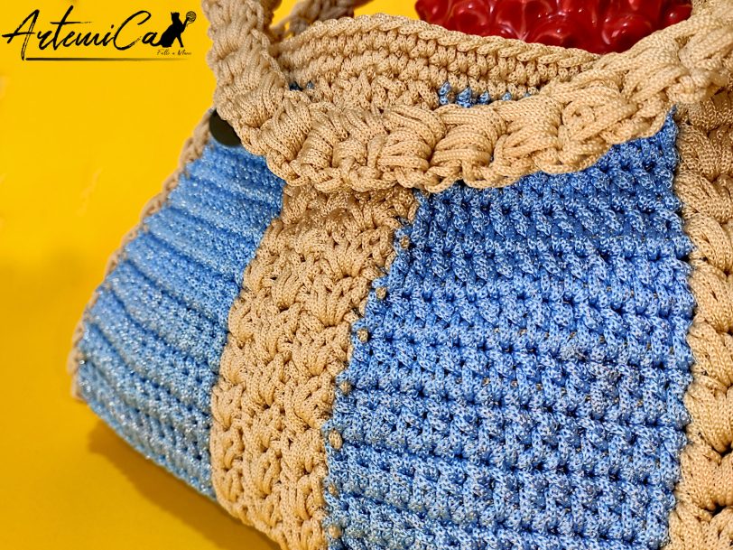 L'immagine mostra una borsa azzurra all'uncinetto con strisce verticali gialle, dotata di un manico coordinato e una chiusura magnetica. Sogno Mediterraneo: il fascino del mare nella tua mano.